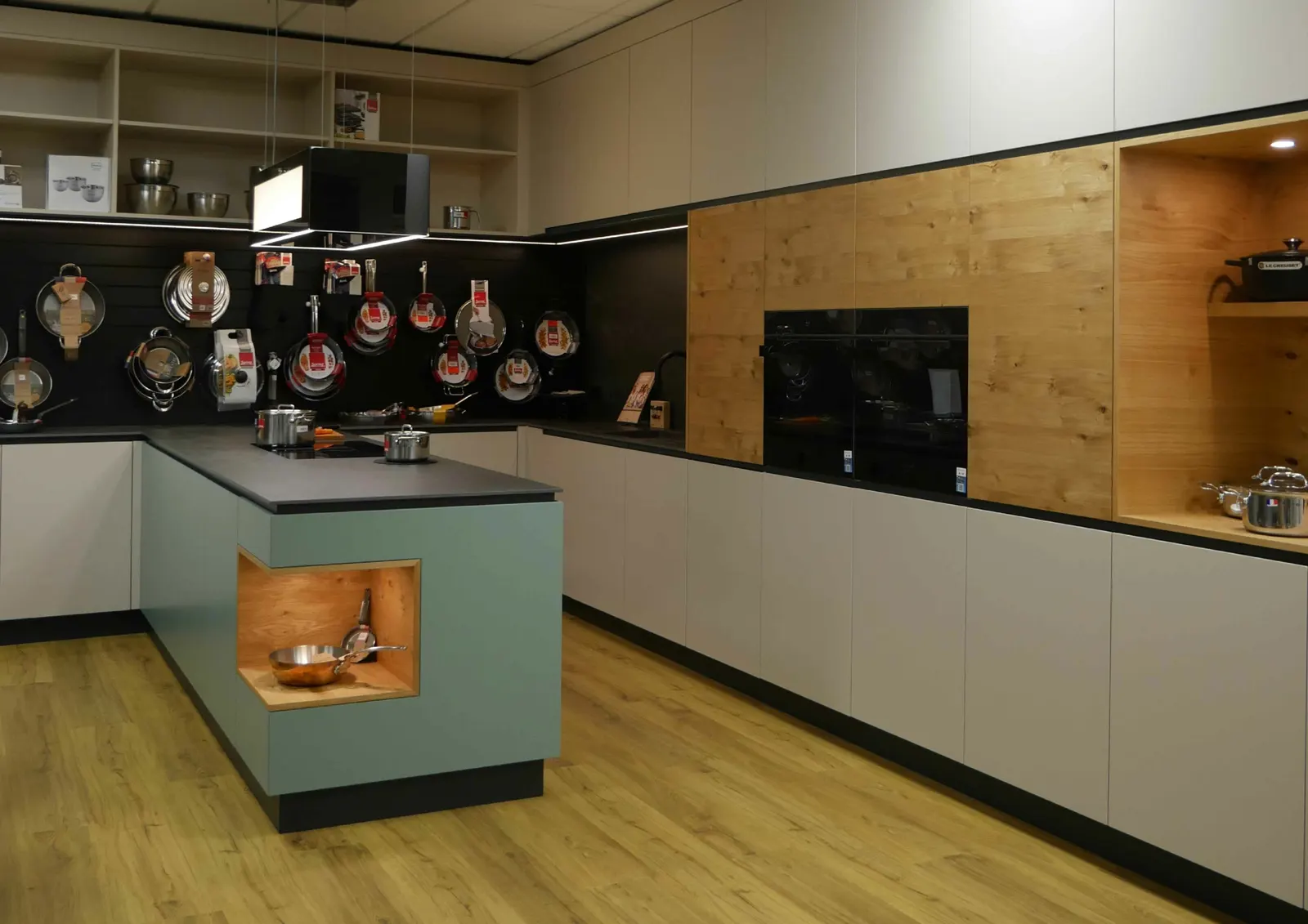 Eine große Küche in einem Ausstellungsraum. Der Küchenblock ist in mintgrün, mit Eckausschnitt aus Holz, der etwas Stauraum bietet. Die restliche Küchenfront ist in weiß, mit Holzelementen. Im Hintergrund befinden sich Töpfe und Pfannen.