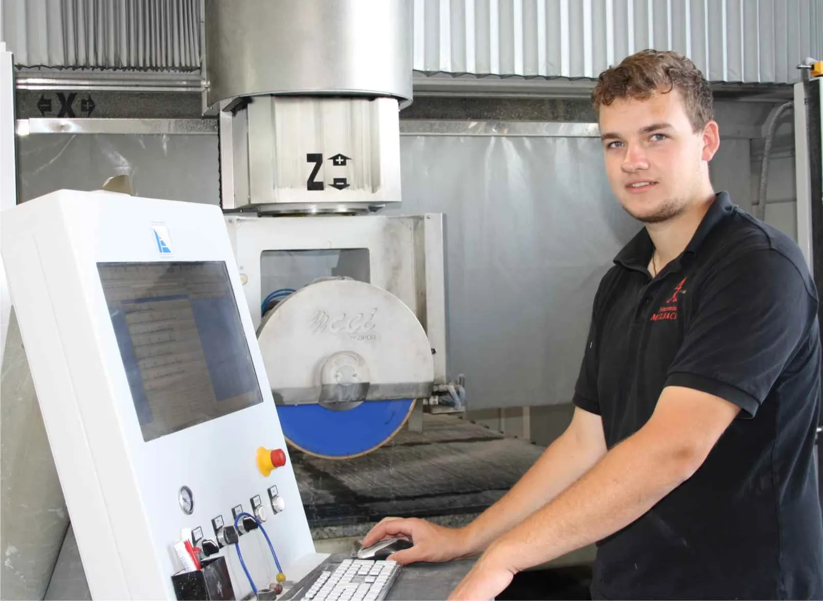 Tobias-Peter Mulbach, Produktionsleiter Naturwerksteinmechaniker, Fachrichtung CNC Steinmetz-&Bildhauermeister. Ein junger freundlicher Mann der eine Maschine bedient. Er hat kurze, braune und lockige Haare, er trägt ein schwarzes Shirt mit dem "Mulbach" Logo.
